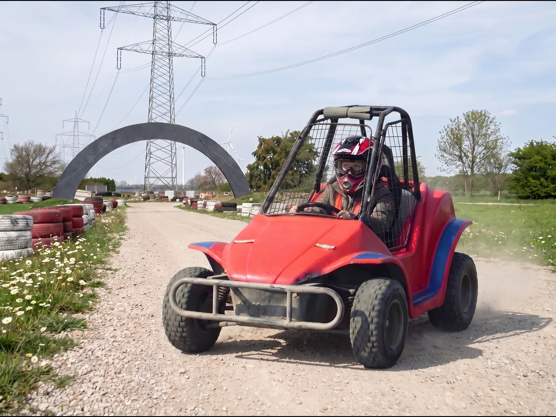 Kind mit Helm fährt im roten Buggy mit Käfig über die Schotterstrecke, im Hintergrund der Torbogen
