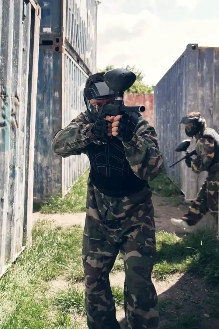 Paintball-Spieler in Tarnkleidung zielt zwischen den Containern in die Kamera